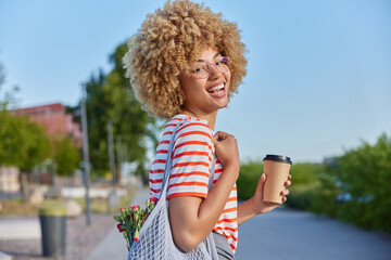Outdoor shot of pleasant looking young woman with curly hair holds paper cup of coffee wears...