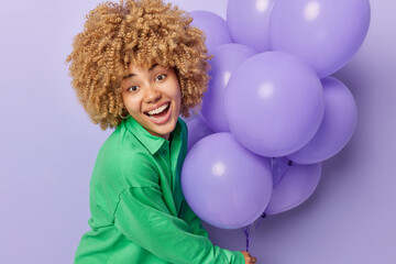 Fototapeta na wymiar Festive event concept. Pretty blonde curly woman dressed in green sweatshirt holds bunch of inflated balloons celebrates special occasion isolated over purple background going to have party.