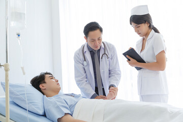 Senior doctor and young male patient who lie on the bed while checking pulse, consult and explain with nurse taking note and supporting in hospital wards.