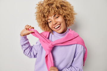 Positive happy woman ties sweater dressed in casual purple sweatshirt smiles broadly has glad expression isolated over grey background. Beautiful female model dresses up and going to have walk