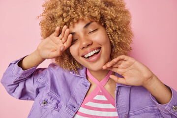Happy dreamy woman with curly blonde hair smiles broadly keeps eyes closed touches face gently recalls pleasant memories enjoys life wears fashionable purple jacket isolated over pink background