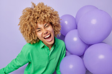 Overjoyed curly haired woman exclaims loudly feels very happy wears green shirt holds bunch of inflated balloons celebrates special occasion enjoys party time isolated over purple background