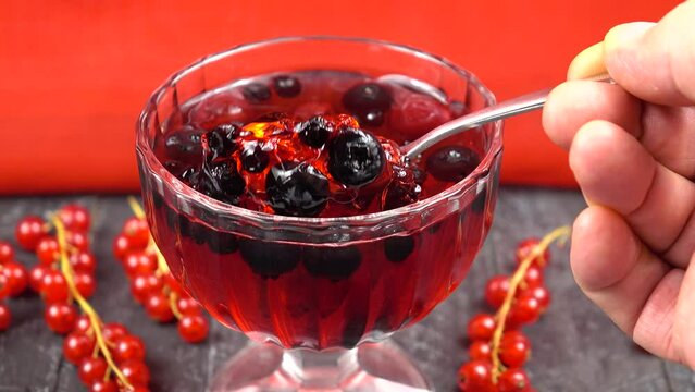 Berry jelly is taken with a spoon from cup standing on a wooden table against a red background 