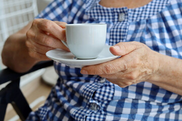 Elderly woman in checkered blue dress sitting with white cup of coffee in wrinkled hands. Concept of fresh morning, energy and enjoying life at retirement