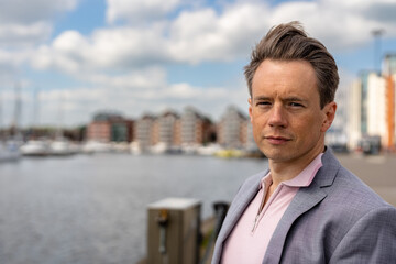 A handsome businessman in a casual grey suit in front of a busy marina with luxury yachts moored up