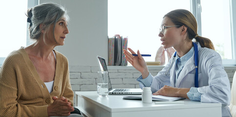 Confident female doctor explaining something to patient while sitting at the desk in medical office