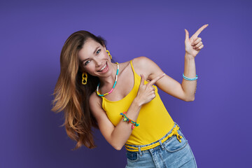 Happy young woman pointing away while standing against purple background