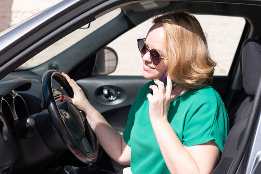 A Smiling Girl In Sunglasses Talking On Phone With Friends, Gesturing While Sitting In The Car. Beautiful Woman Using Mobile Phone Driving Car. Girl Talking To The Phone In Her Car Holding The Wheel