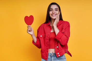 Happy young woman holding paper heart while standing against yellow background