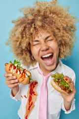 Photo of curly haired woman with curly hair holds delicious hot dog and burger enjoys eating fast...