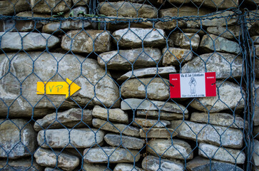 La freccia gialla della Via Postumia e il logo della Via del Volto Santo legati ad una rete che core un muro di pietre lungo la Via degli abati, cammino che parte da Pavia e arriva a Pontremoli © Andrea Vismara