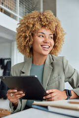 Successful positive woman with curly hair readis financial news on internet websites holds digital tablet dressed in formal clothes sits against cozy interior looks away with positive smile on face