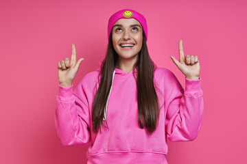 Beautiful young woman in funky hat pointing up and smiling against pink background
