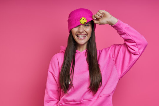Happy young woman in hooded shirt adjusting her funky hat against pink background