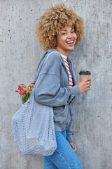 Vertical shot of positive curly haired woman dressed in sweatshirt and jeans holds takeaway coffee...