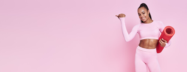 Happy woman in sports clothing carrying exercise mat and pointing away against pink background