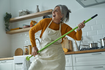 Playful senior woman holding mop like guitar while standing at the domestic kitchen