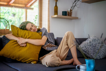 Cheerful curly cute young woman sitting at home on gray sofa hugging yellow pillow, relaxing at home concept