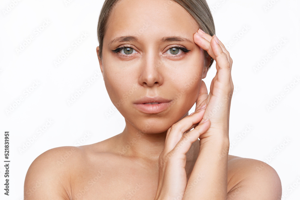 Wall mural Portrait of a young attractive tanned caucasian blonde woman with makeup touching the skin of her face isolated on a white background. Beauty and fashion. Skin care