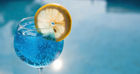 A glass of delicious blue cocktail "Blue Lagoon" and yellow lemons on the background of the pool. Alcoholic cocktail juicy fruit blue with curacao liqueur, ice cubes and a slice of lemon. 