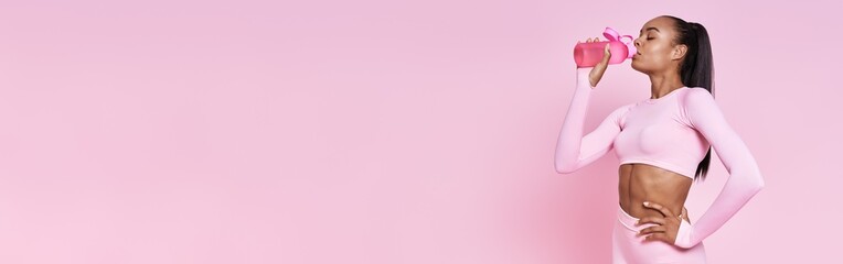 Beautiful African woman in sports clothing drinking water against pink background © gstockstudio