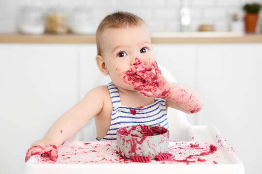 Funny baby in high chair with messy face licking his fingers