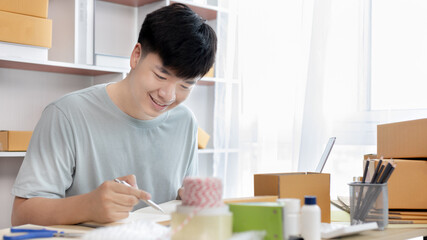 Young Asian man is writing down the customer's details and addresses on the notebook or box in order to prepare for shipping according to the information, Packing box, Sell online, freelance working.