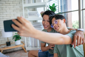 Happy Asian gay couple use mobile phone video call online or using an app on sofa, LGBTQ concept.
