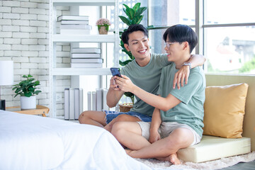 Happy Asian gay couple use mobile phone video call online or using an app on sofa, LGBTQ concept.