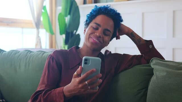 Young positive African American woman making video call via mobile phone waving hand in greeting and telling friends how week went sits on couch indoors. Technology, Applications, Internet