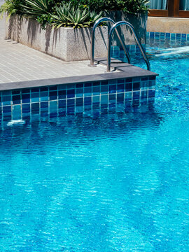 Poolside decoration with green leaves in the stone block near the ladder at the swimming pool with copy space, vertical style. Empty outdoor pool on sunshine day in summer.