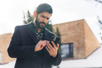 Hispanic bearded businessman wearing coat outside on winter morning swiping through messages on...
