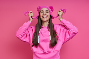 Happy young woman in hooded shirt playing with her funky hat against pink background
