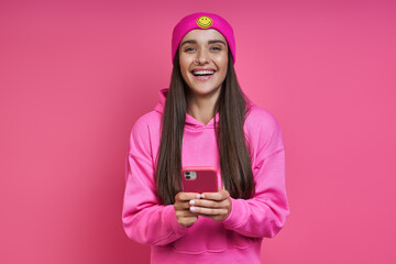 Beautiful young woman in hooded shirt and funky hat holding smart phone against pink background