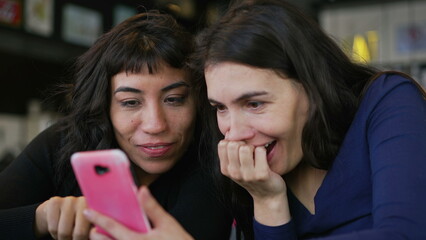 Two young women looking at smartphone device screen smiling. Female friends sharing information watching content online