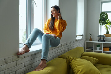 Dreamful young woman looking through a window while sitting on the window sill at home