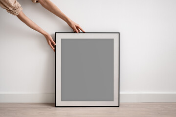 Female hands showing and holding big black frame with empty space on white wall background. Minimalist blank mockup template to display picture, poster, art, design, certificate with copyspace around.