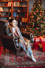 Woman Sitting in an Armchair with Cup in her Hands. Interior of House Decorated in New Year's Decor.