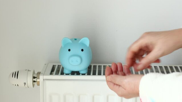 Piggy bank standing on the radiator, Woman in a winter bathrobe, puts money into the piggy bank, Creative concept, High costs of heating the house, Problems with energy supply