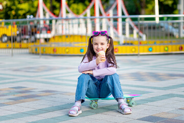 portrait of a child girl in an amusement park in summer eating ice cream sitting on a skateboard...