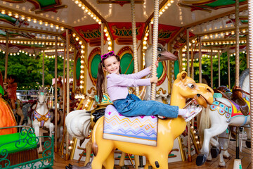 a child girl in an amusement park rides on a carousel and smiles with happiness, the concept of weekends and school holidays - obrazy, fototapety, plakaty