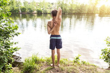 Young man at the lake shore doing exercises before swmming into the water in sunlight. Sport, yoga, fitness outdoors in nature, healthy lifestyle