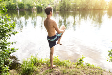 Young man at the lake shore doing exercises before swmming into the water in sunlight. Sport, yoga, fitness outdoors in nature, healthy lifestyle