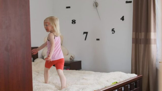 Blonde little girl indulges and runs around room on bed alone. Kid with dirty painted face.