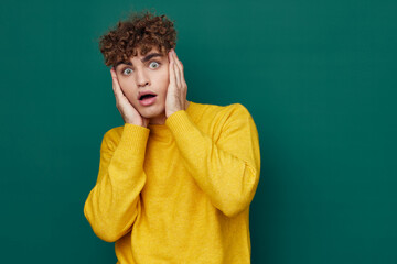 Obraz na płótnie Canvas a shocked man in a yellow sweater stands on a green background and holds his head with his hand, his mouth wide open, looking into the camera from shock