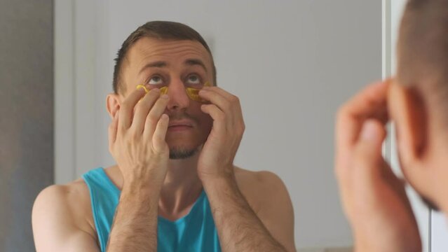 Brunet man puts patches under eyes and massages his fingers around eye area. Guy touched by his reflection in mirror.