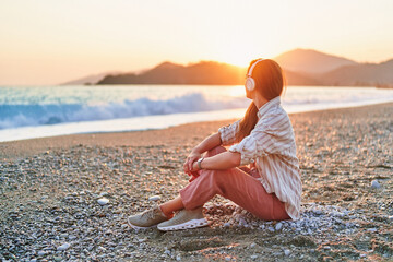 Calm woman sitting alone and listening relax traveling music on the seashore at sunset time....