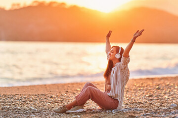Fototapeta na wymiar Happy alone woman with open arms and closed eyes enjoys of listening relax traveling music on the seashore at sunset time