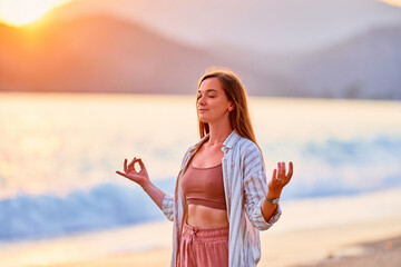 Portrait of calm serene peaceful woman with closed eyes in lotus position meditates and enjoys beautiful moment life on the seashore at sunset time - obrazy, fototapety, plakaty