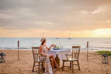 Romantic date on the sunset beach. Woman sitting alone on the table with gourmet food, drinking champagne from glass. Happy woman in luxury outdoor restaurant happy and smiling after proposal